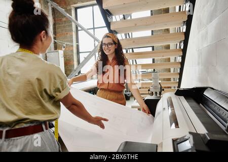 Two young fashion designers standing under staircase in studio or workshop and printing sketches of new clothing items together Stock Photo