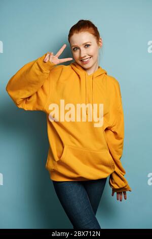 Redhead girl in a yellow oversized sweatshirt stands on a blue background and smiling shows a peace sign. Stock Photo