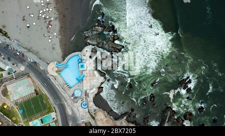 Aerial photo of an outdoor swimming pool at the coastline next to the rocks and crashing waves. Stock Photo