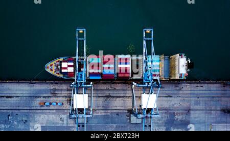 Aerial top view of the boat with its containers being unloaded with the crane, at the container port, terminal. Stock Photo