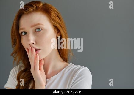Beautiful red-haired girl with clean skin, blue eyes and freckles in a white T-shirt, looks into the frame, covering lips with fingers Stock Photo