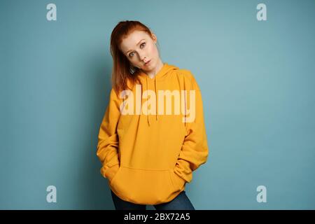 A red-haired girl in a yellow oversized sweatshirt stands on a blue background, bowing her head and putting hands in pockets Stock Photo