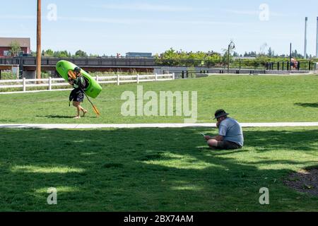 Two people at McKay Park in downtown Bend Oregon. One man is reading a book, the other is taking his kayak to whitewater portion of park. Stock Photo