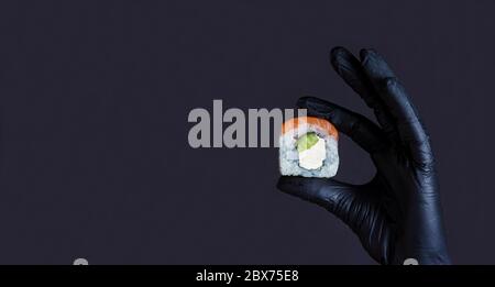 Diet food concept. Female hand in rubber gloves holding sushi salmon roll on a dark background with copyspace. Sushi menu. Japanese food. Stock Photo