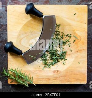 Chopping fresh rosemary with mezzaluna curved knife on board, top view on slate. Stock Photo