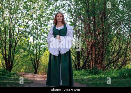 Beautiful medieval young woman in traditional historical female green and white costume at nature. Fantasy girl in long dress or gown walking at path Stock Photo