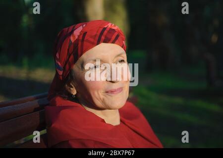 Female portrait of mature woman dressed in red clothes and sitting on the bench and enjoying a good weather and sunset in the park during a spring or Stock Photo