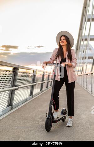 A young beautiful girl is standing next to her electric scooter with one foot on a standing platform. She is wearing a hat and she is business-trained Stock Photo