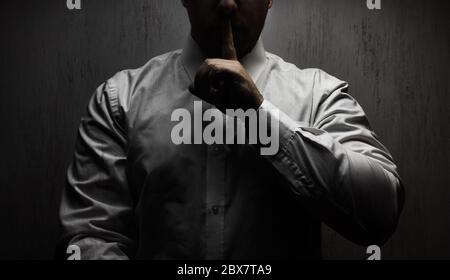 Photo of a scary horror man in white shirt standing under upper light on black background showing silence hand sign. Stock Photo