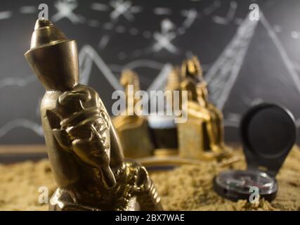 Ancient old pharaoh closeup metal statuettes standing on a sand surface with a blackboard drawn background & compass. Stock Photo