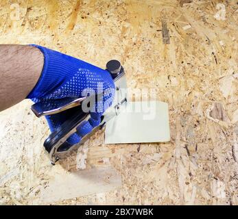 Worker hand in blue dotted gloves holding steel industrial stapler and nailing memo paper on a wooden surface board. Stock Photo