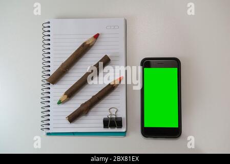 Top view of office desk with blank note book, rustic colored pencils and smartphone with green screen. Close up to insert image on cell phone screen. Stock Photo