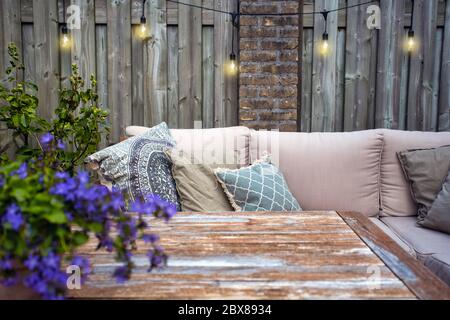 Stylish outdoor garden furniture, sofa with cushions and lamps, light bulbs hanging, cozy modern corner on terrace Stock Photo