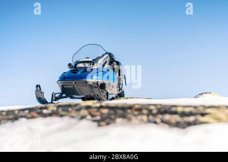 One blue snowmobile stands higher up on snow with stones in Norwegian mountains, warm day and very bright sun, no clouds, snow starting to smelt, norw Stock Photo
