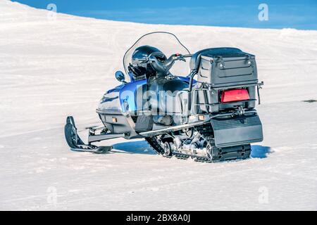 One blue snowmobile stands on a crust snow at slope in Norwegian mountains, back view. Warm day and very bright sun, no clouds, mountains winter lands Stock Photo