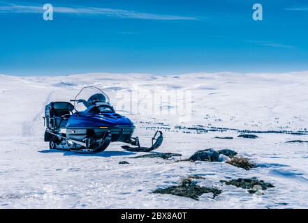 One blue snowmobile stands on a crust snow in Norwegian mountains, warm day and very bright sun, no clouds, snow starting to smelt, snowmobile road, v Stock Photo