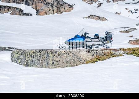 One blue snowmobile stands on snow close to stones in Norwegian mountains, warm day and bright sun, snow smelts, norwegian mountains landscape, side v Stock Photo