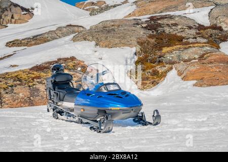 One blue snowmobile stands on snow close to stones in Norwegian mountains, warm day and bright sun, snow smelts, norwegian mountains landscape, front Stock Photo