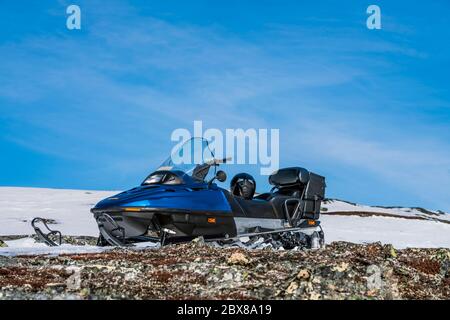 One blue snowmobile stands higher up on snow with stones in Norwegian mountains, warm day and very bright sun, no clouds, snow starting to smelt, norw Stock Photo
