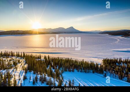 AERIAL: Sunset golden light over Norwegian snowy winter mountains and Rossvatnet lake, pine and birch trees, early spring, calm blue skies. Hattfjelld Stock Photo