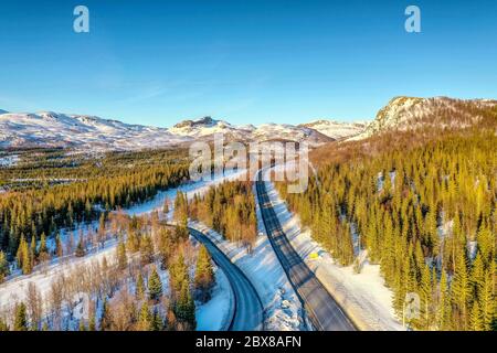 AERIAL: Sunset light over Norwegian snowy winter mountains with birche and pine trees, two roads, early spring, calm blue skies. Rossvatnet lake area,