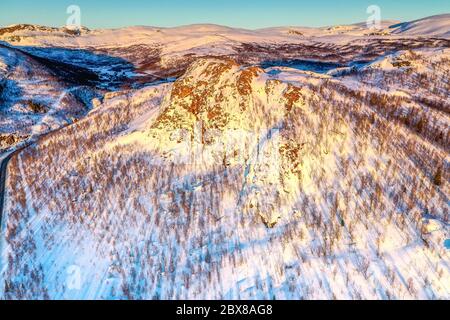 AERIAL: Sunset golden light over Norwegian snowy winter mountains with birch and pine trees, early spring, calm blue skies. Rossvatnet lake area, Hat Stock Photo