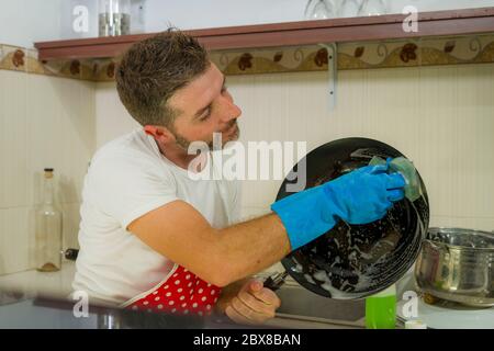 home lifestyle portrait of happy and attractive man in red apron smiling at kitchen washing dishes and cooking pan cheerful doing housework and domest Stock Photo