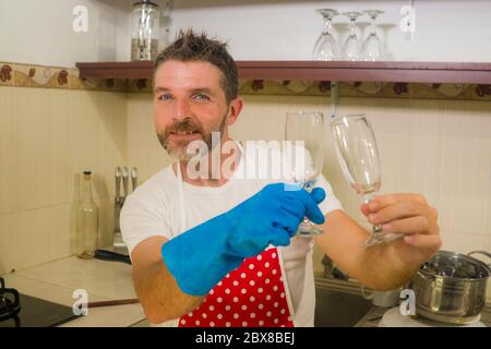 home lifestyle portrait of happy and attractive man in red apron smiling at kitchen washing dishes and wine glass doing housework and domestic chores Stock Photo