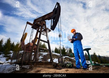 Male worker using notebook controlling work of petroleum pump jack. Operator in work overalls and helmet standing near oil pumping unit. Concept of petroleum industry and oil extraction. Stock Photo