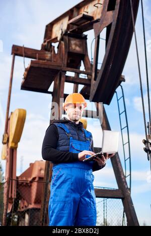 Vertical portrait of oil man, wearing blue overalls and orange helmet, with a laptop, standing with his back to an oil rig, checking oil pumping unit, making notes in his computer Stock Photo