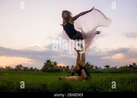 outdoors sunset acroyoga workout - young happy and fit couple practicing acro yoga drill at beautiful rice field enjoying nature doing acrobatic pose Stock Photo