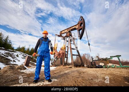Oil worker during his working day in oil field, man wearing blue overalls and orange helmet, holding pipe wrench, looking at camera under beautiful sky. Concept of oil extraction, petroleum industry. Stock Photo