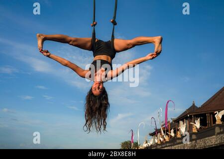 aero yoga workout - young attractive and athletic woman practicing aerial yoga exercise training acrobatic  body postures isolated on blue sky in bala Stock Photo