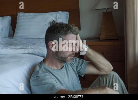 dramatic home portrait of young depressed and desperate man sitting on bedroom floor next to bed suffering depression and anxiety feeling overwhelmed Stock Photo