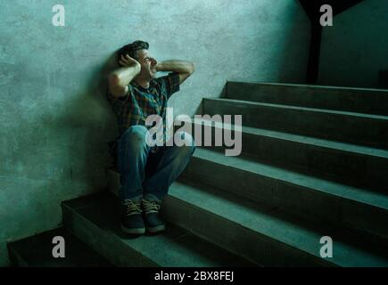 dramatic portrait of young depressed and paranoid man sitting outdoors on dark grunge street corner staircase feeling sick suffering depression proble Stock Photo