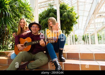 Two girls smile and enjoy themselves while a musician plays a guitar sitting on a staircase with a bright background of trees Stock Photo