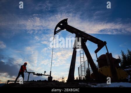 Silhouette of oilman working with an oil pump jack in oilfield. Worker tightening the bolt with gas wrench, against beautiful evening sky, low angle view. Concept of petroleum industry, oil extraction Stock Photo