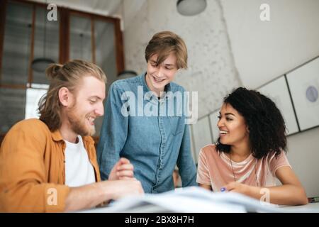 Portrait of young cheerful people discussing something in office. Two boys with blond hair and girl with dark curly hair working together on new proje Stock Photo