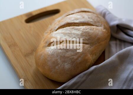 A loaf of homemade baked sourdough bread with three parallel cuts scoring on a wooden cutting board with a kitchen towel Stock Photo