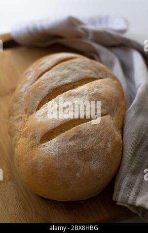 A loaf of homemade baked sourdough bread with three parallel cuts scoring on a wooden cutting board with a kitchen towel Stock Photo