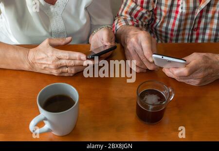 Top view of old people using smartphones at home while enjoying cups of hot drinks. Concept of elderly people and technology. Stock Photo