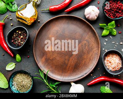 Empty wooden plate and frame of spices, herbs and vegetables on a dark stone background. Top view, flat lay. Stock Photo