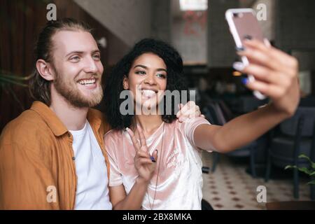 Beautiful young couple making photos on frontal cellphone camera in restaurant. Pretty African American lady making photo with friend in cafe. Friends Stock Photo