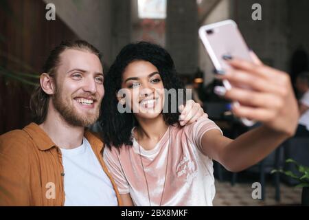Joyful young couple making photos on frontal cellphone camera in restaurant. Nice African American lady making photo with friend in cafe. Smiling frie Stock Photo