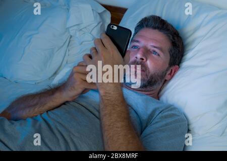 dim light night shot of young attractive and happy man with blue eyes lying on bed late networking late on mobile phone tired and sleepy texting onlin Stock Photo