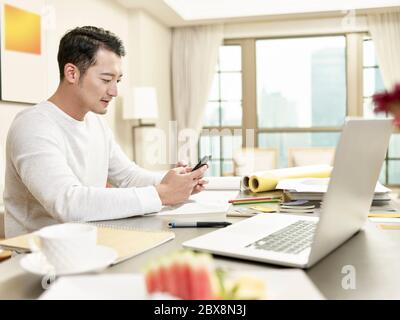 young asian man design professional working from home sitting at kitchen using cellphone (artwork in background digitally altered) Stock Photo