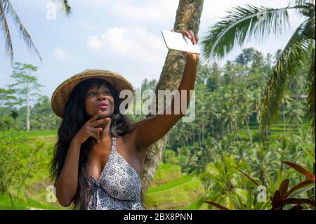 young attractive happy afro american black woman tourist taking selfie portrait photo with mobile phone camera while exploring rice fields forest and Stock Photo
