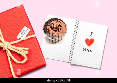 valentines day card background with red present gift box isolated on pink table next to chocolate cupcake and notepad with I love you handwritten in r Stock Photo