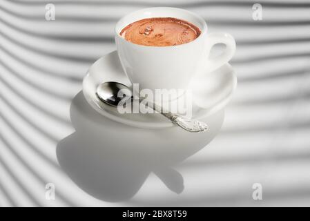 White cup of coffee on white background with shadows from the window blinds. Best start to the day. Copy space Stock Photo
