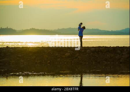 Silhouette of young happy man with his little son or daughter in his arms , the father raising up the baby in front of the sea at beach boardwalk enjo Stock Photo